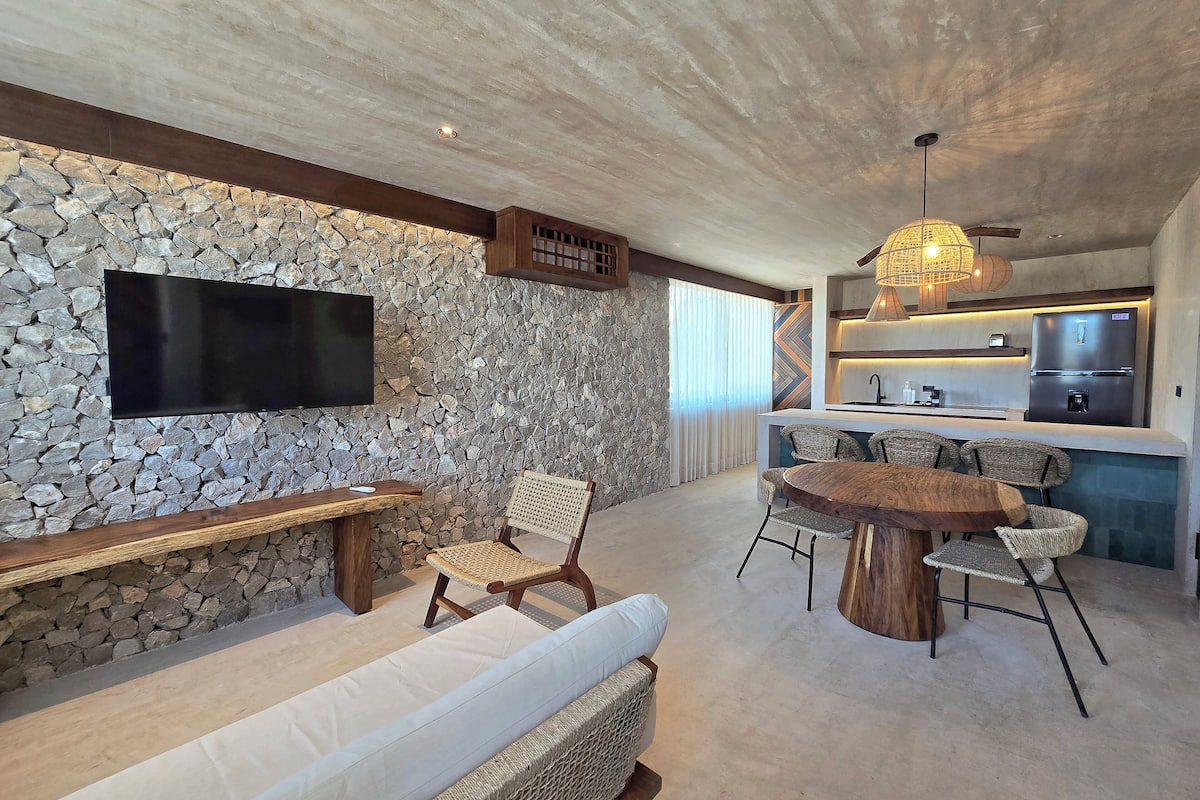 A modern living area with a stone wall, a flat-screen TV, a wooden bench, wicker chairs, a round wooden table, and a kitchen area with a refrigerator and sink.