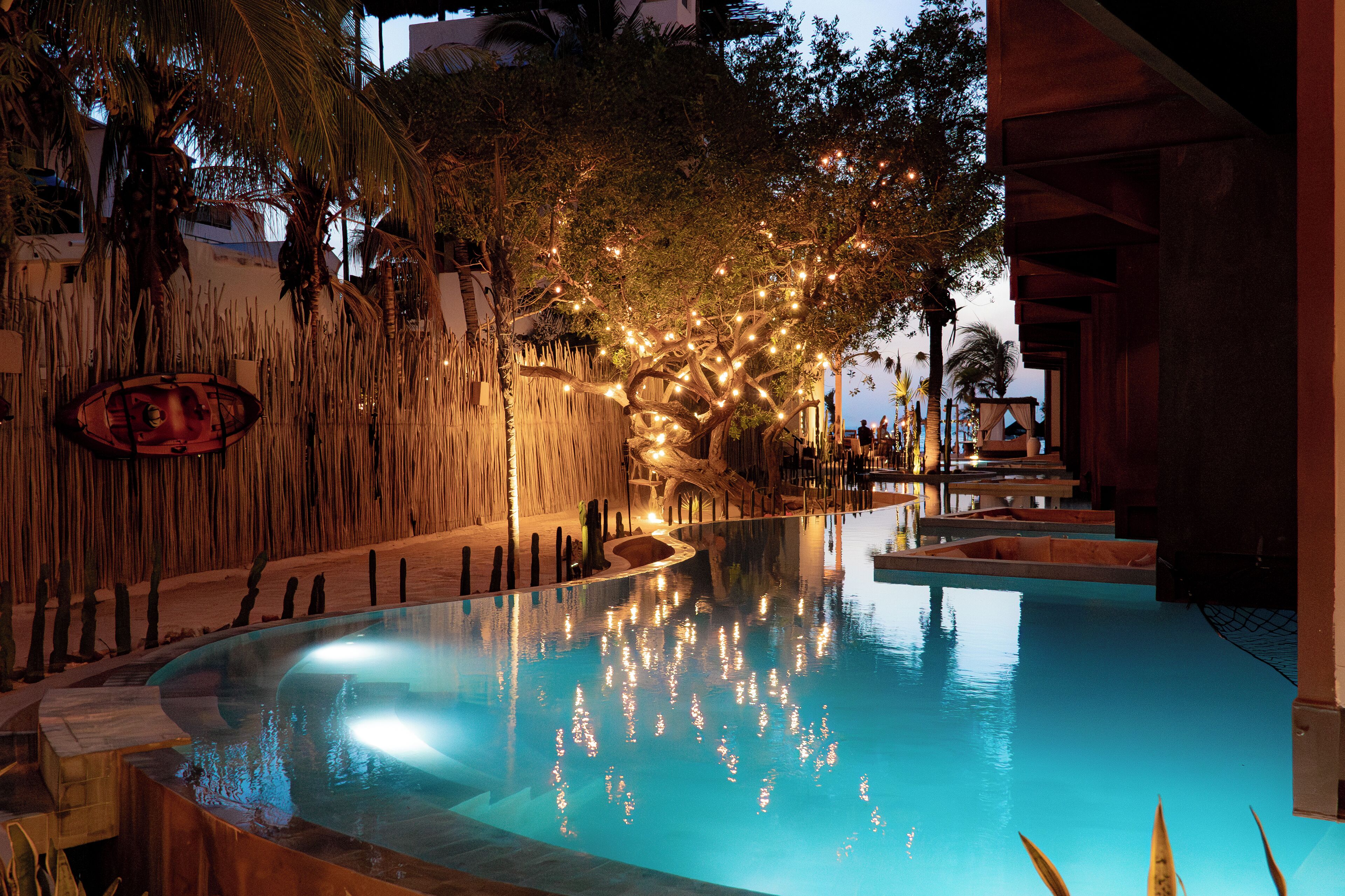 Una piscina con una pared de bambú y luces colgantes, rodeada de palmeras y un edificio.