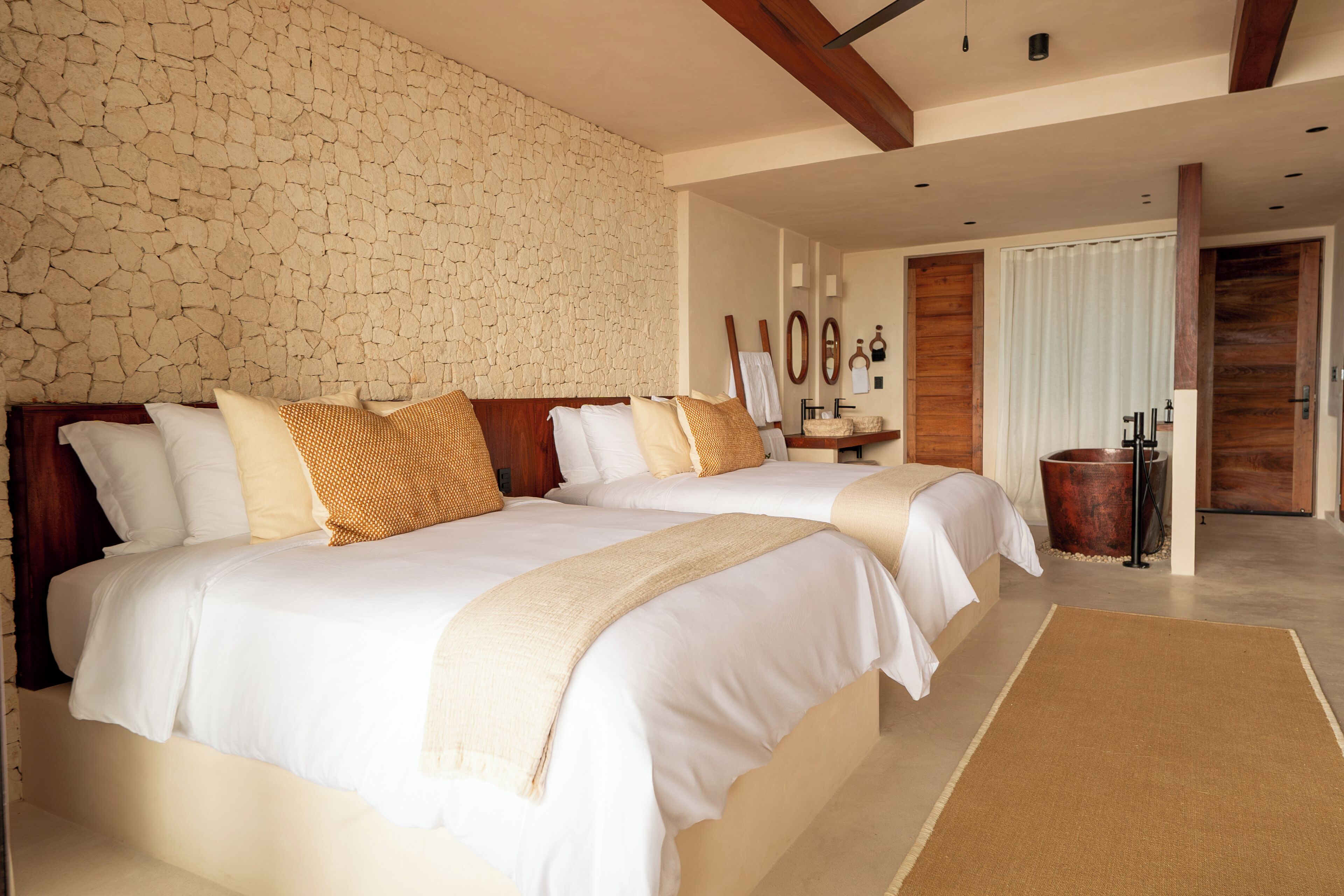 A bedroom with two beds, a stone wall, wooden beams, a mirror, and a bathroom with a copper sink.