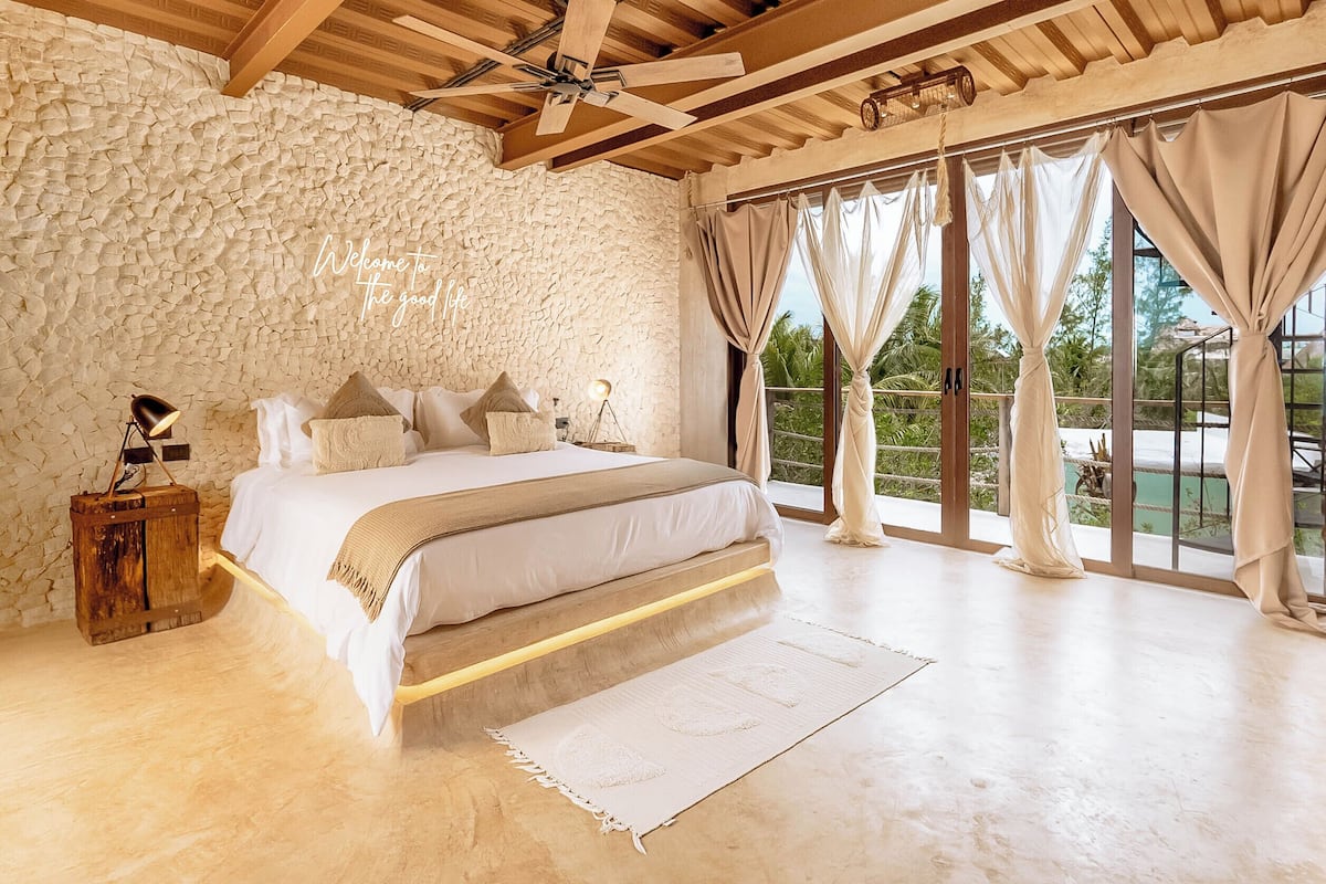 A bedroom with a stone wall, a bed with white bedding, a wooden headboard, and a view of greenery through large windows.