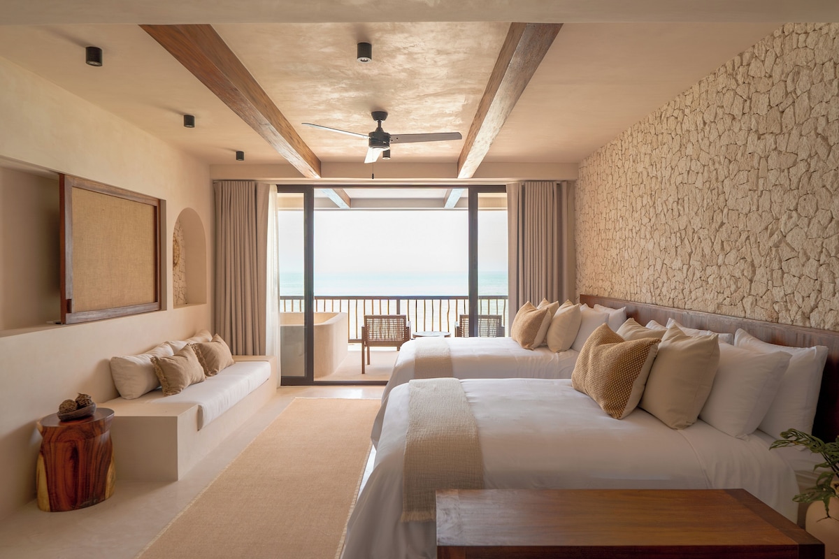 Habitación de hotel con una cama grande, un sofá más pequeño, una mesa de centro de madera y vista al océano desde un balcón.