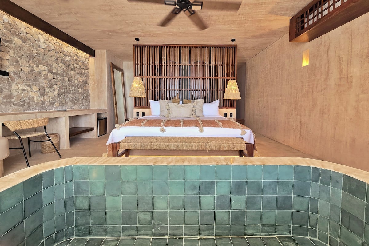 A bedroom with a large bed, a wooden headboard, a stone wall, a desk with a chair, and a bathtub with green tiles.