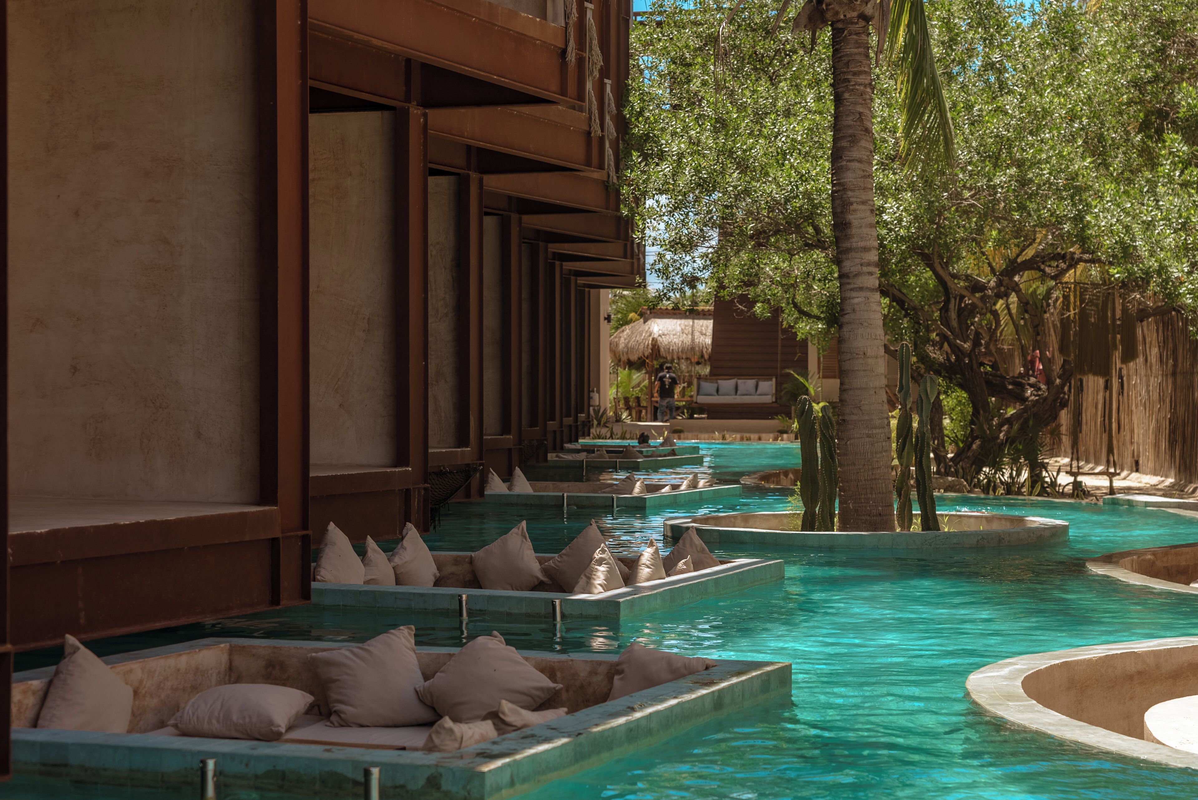 A series of rectangular water features with seating, surrounded by a wooden structure and palm trees.