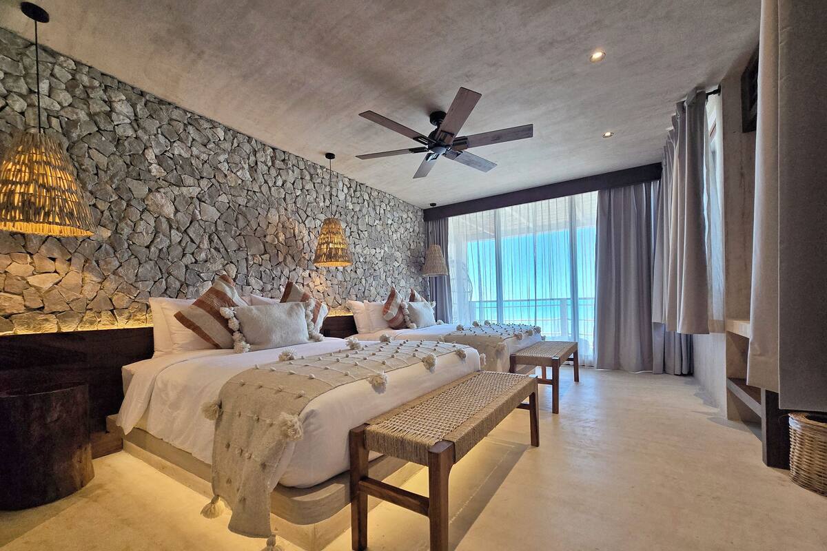 A spacious bedroom with a stone wall, a large bed with multiple pillows, a ceiling fan, and a view of the outdoors.