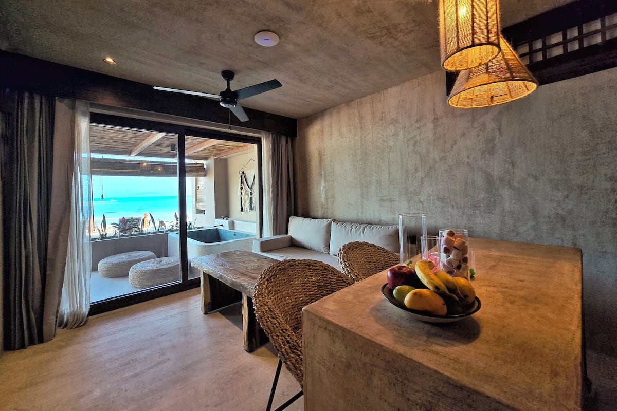 A room with a view of the ocean, a ceiling fan, a table with fruit, and a large window.