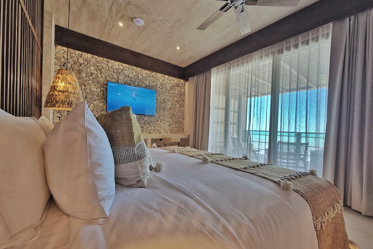 A spacious bedroom with a large bed, a stone wall, a ceiling fan, and a TV mounted on the wall.