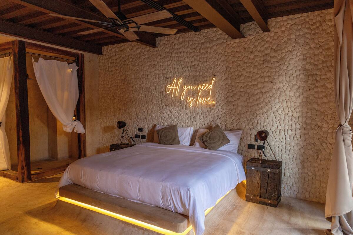 A bedroom with a stone wall, a bed with white bedding, and a neon sign that says "All you need is love."