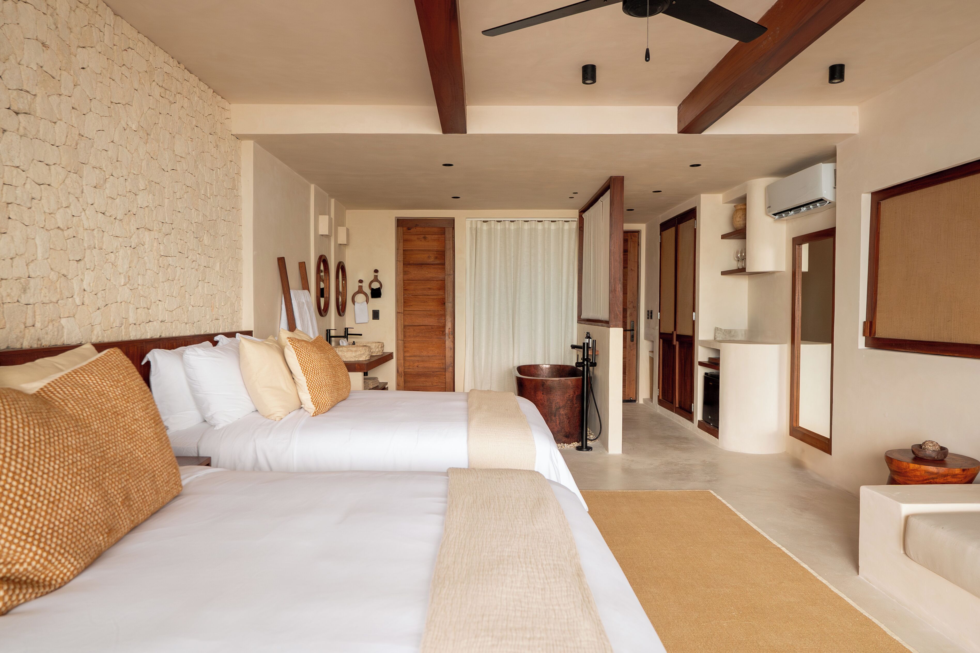 A hotel room with two beds, a stone wall, wooden beams, a ceiling fan, a bathroom with a bathtub, and a large mirror.