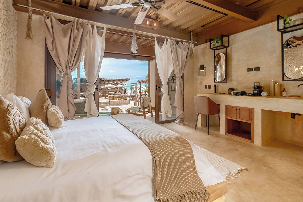 A bedroom with a large bed, a wooden ceiling, a view of the beach, a bathroom with a sink and mirror, and a window with curtains.