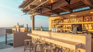 Un bar en la playa con mostrador de madera, taburetes y una variedad de botellas.