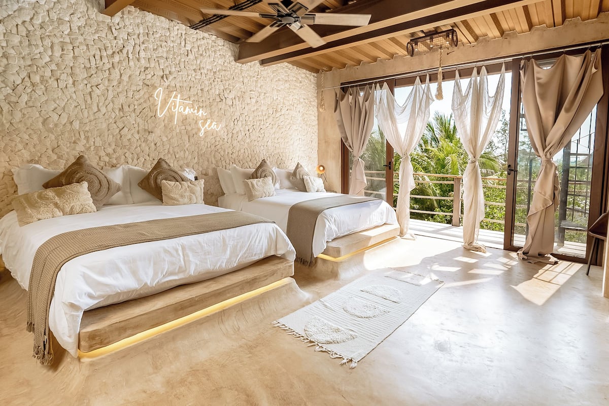 A bedroom with two beds, a stone wall, large windows with curtains, and a ceiling fan.