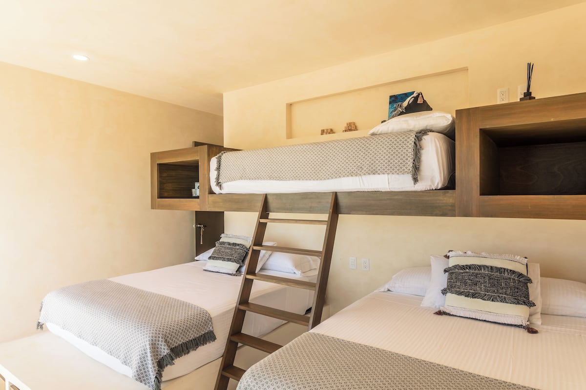 A bunk bed room with a ladder, two beds, and a shelf above the upper bunk.