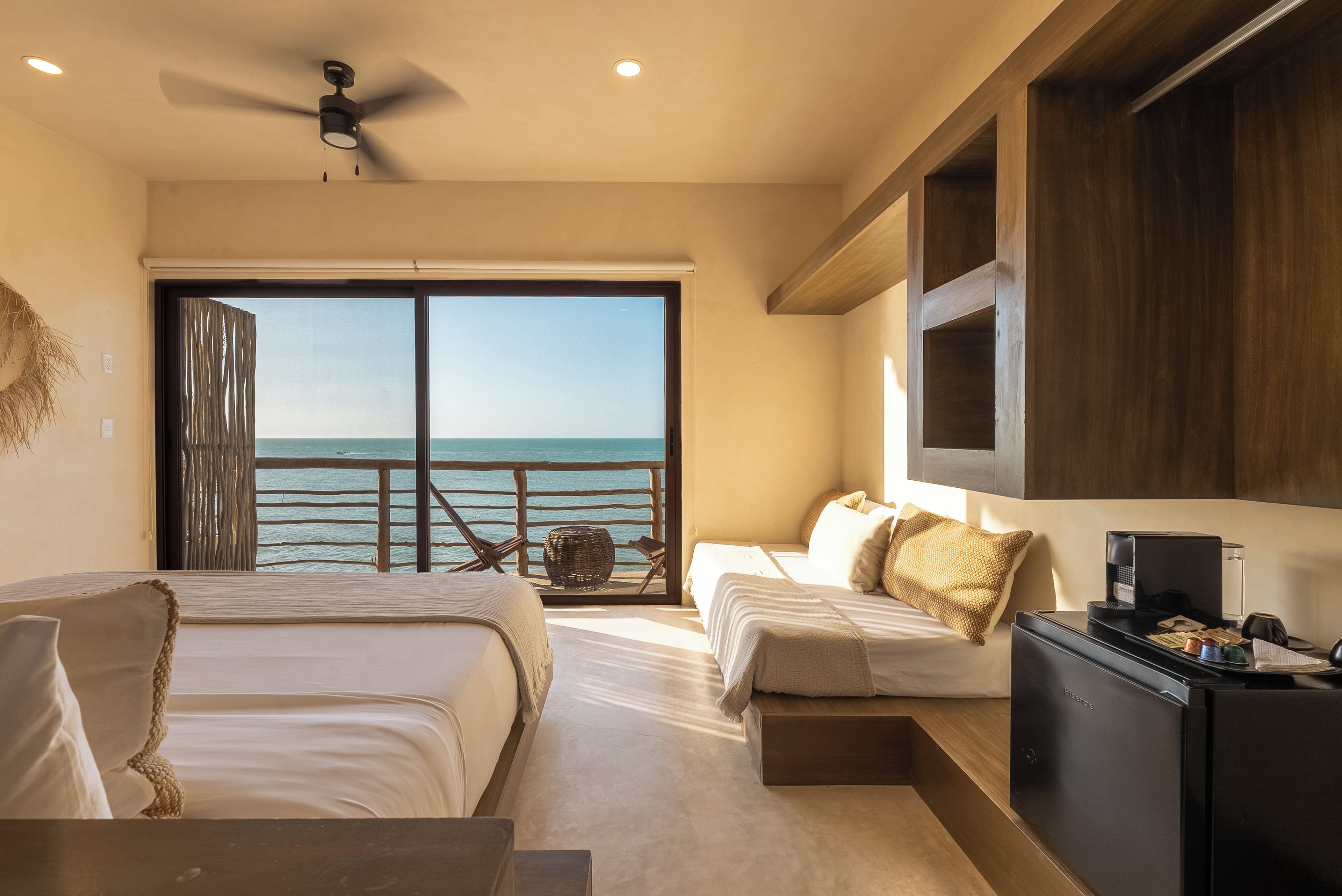 A hotel room with a bed, a desk, a chair, a view of the sea, and a ceiling fan.