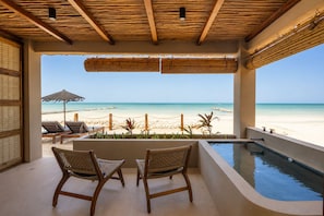 Un balcón con vista a la playa, dos sillones de mimbre, una piscina y un techo de paja.