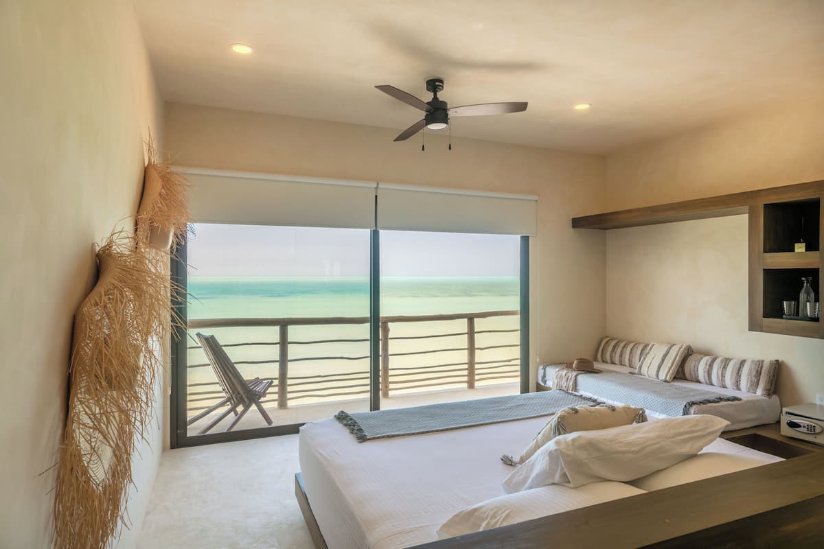 A bedroom with a bed, a bedside table, a ceiling fan, and a view of the beach.