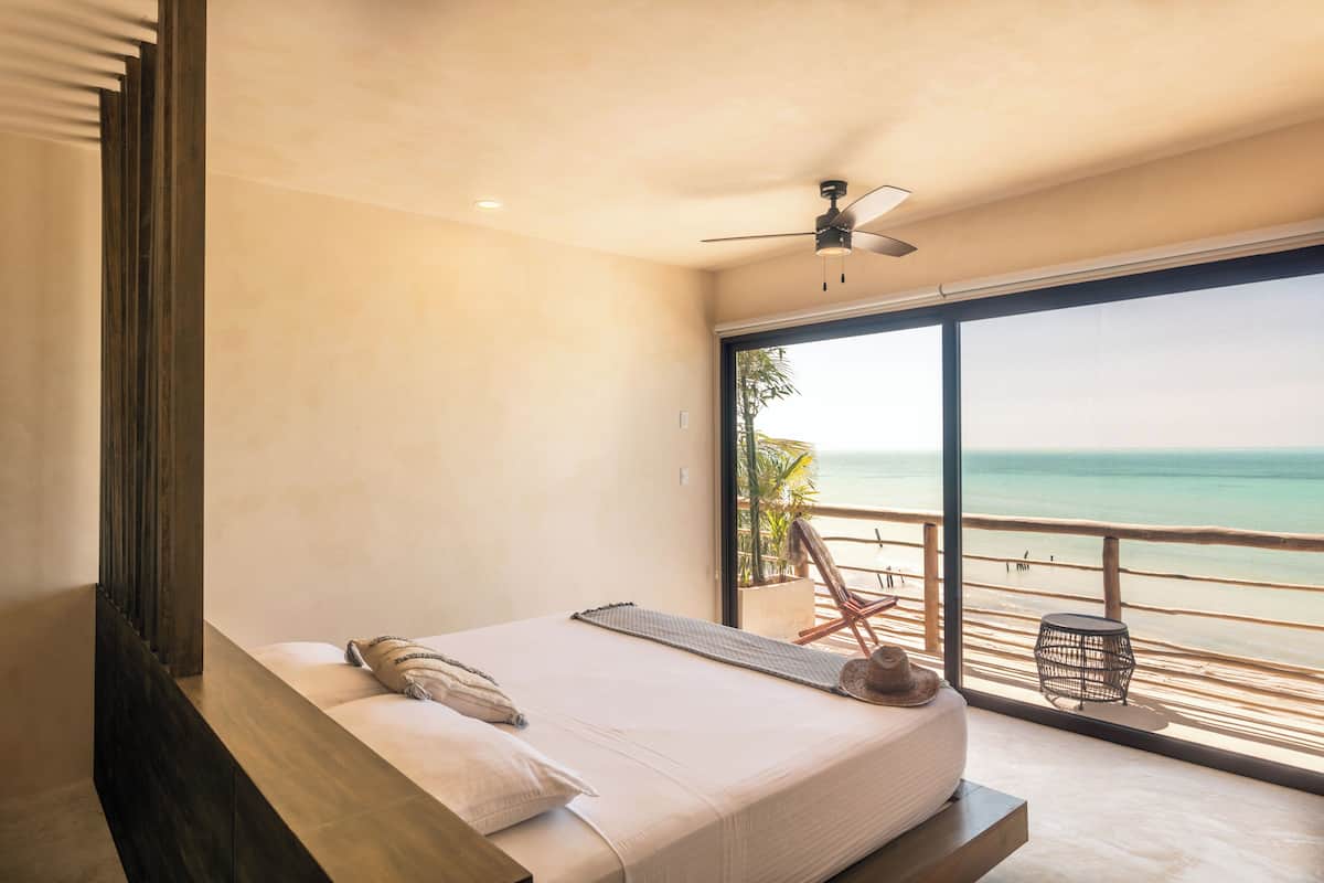 A bedroom with a large bed, a ceiling fan, and a view of the beach through a sliding glass door.