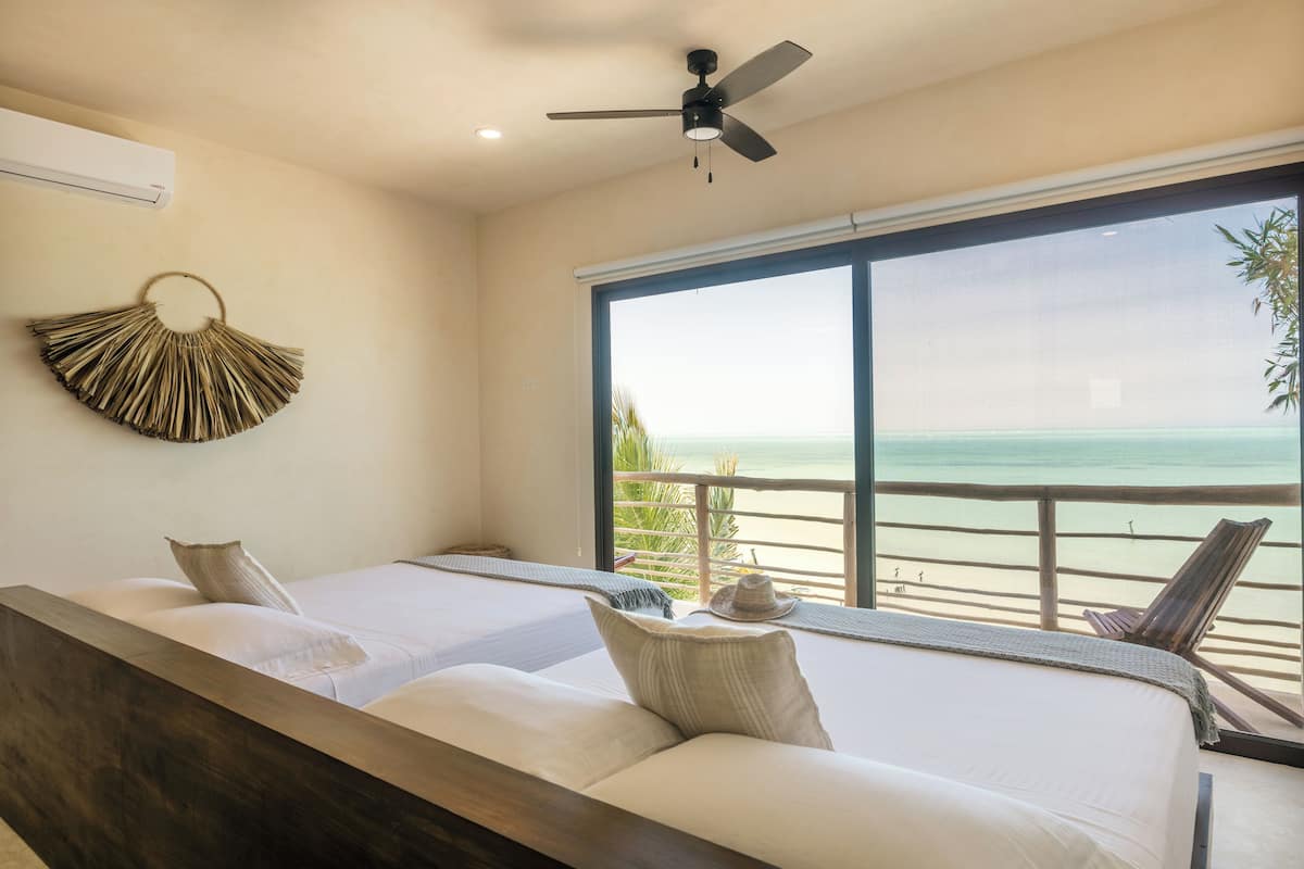 A bedroom with two beds, a ceiling fan, and a large window overlooking the beach.
