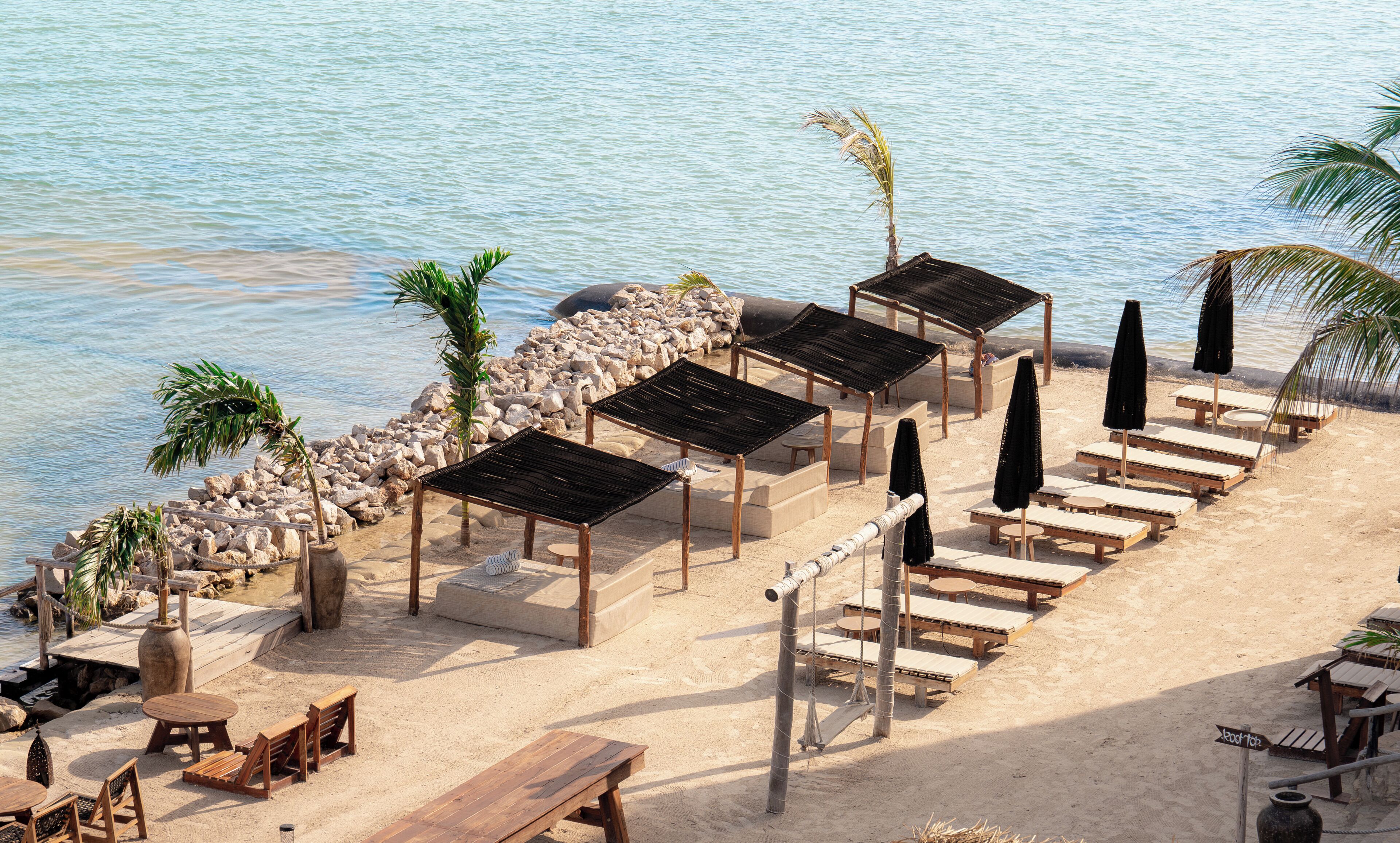 A beachside lounge with wooden tables and chairs, a stone wall, and umbrellas.