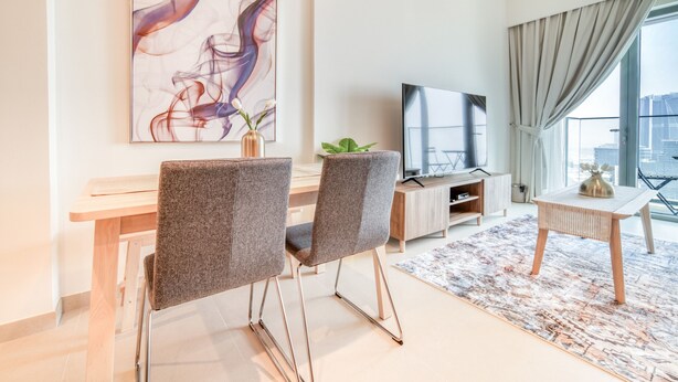 A modern living room with a large abstract painting, a flat-screen TV, a dining area with two chairs, a small table, and a view of the city through large windows.