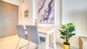 A modern dining area with a white table and two grey chairs, a large abstract painting on the wall, and a potted plant.