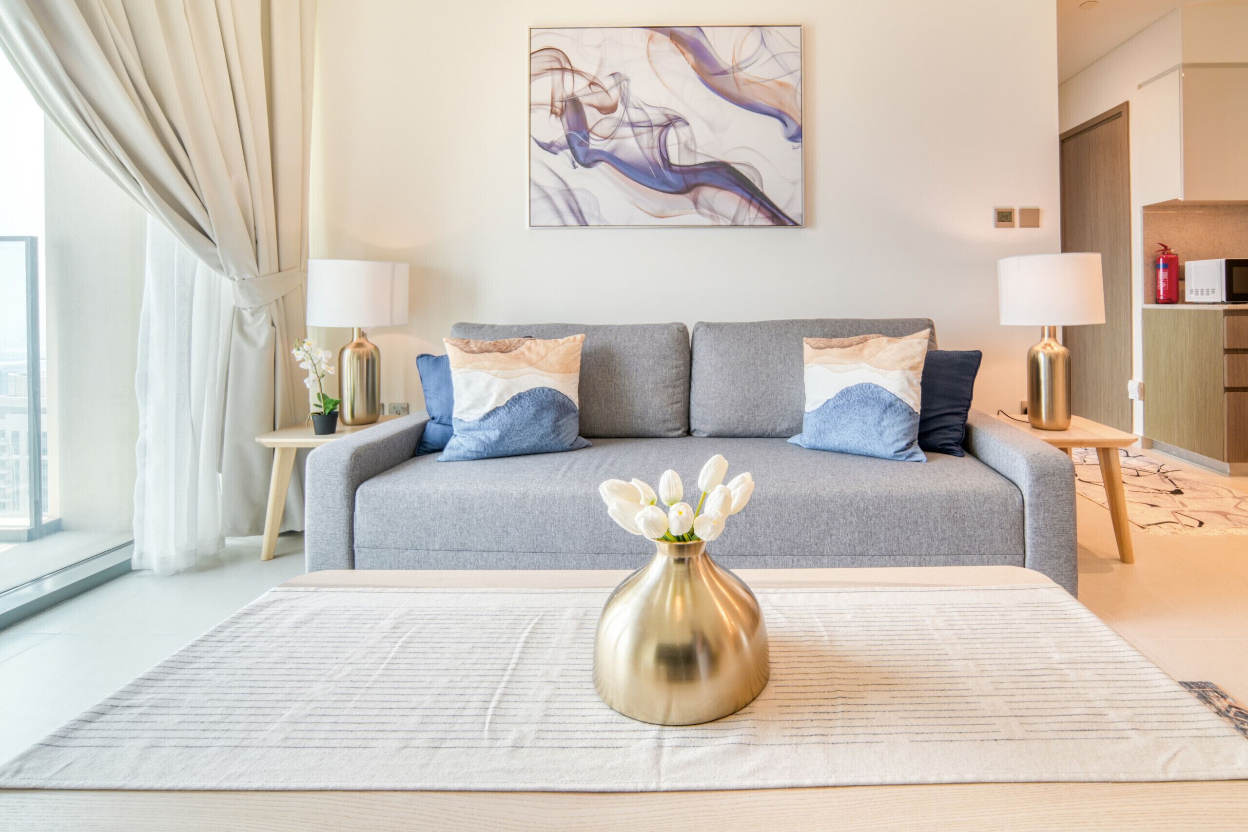 Una sala de estar moderna con un sofá gris, dos mesitas de noche con lámparas, un jarrón con flores blancas y un cuadro en la pared.