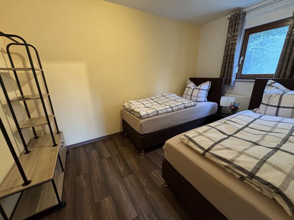 Una habitación de hotel con dos camas, suelo de madera, una estantería metálica y una ventana con cortinas.