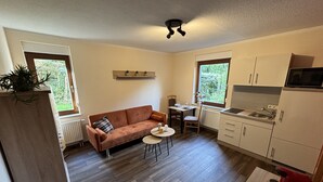 A compact living space with a sofa, a small dining table, a kitchen area with cabinets and a microwave, and large windows providing natural light. - Ferienwohnung Berger (Penig)