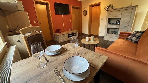 A dining area with a wooden table set for two, a sofa, and a television mounted on the wall. - Ferienwohnung Berger (Penig)
