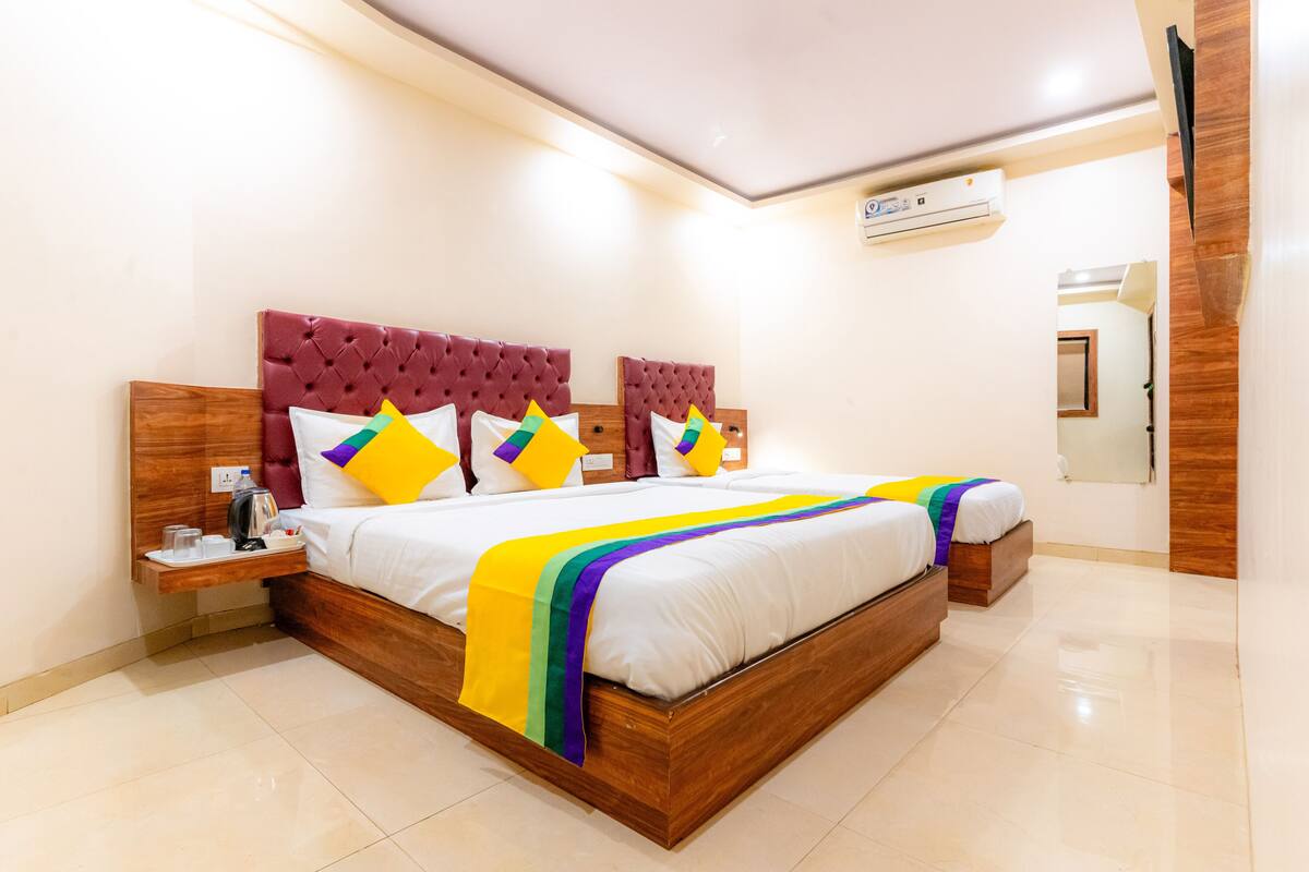 A modern hotel room with a large bed, a wooden headboard, a bedside table with a lamp, and a small shelf with a water bottle and glasses.