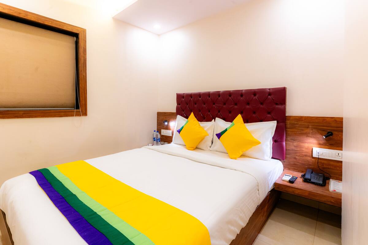 A hotel room with a bed, a nightstand, a red upholstered headboard, and a white wall.