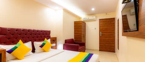 A hotel room with a large bed, a red upholstered headboard, a wooden wardrobe, a red armchair, and a mounted TV.