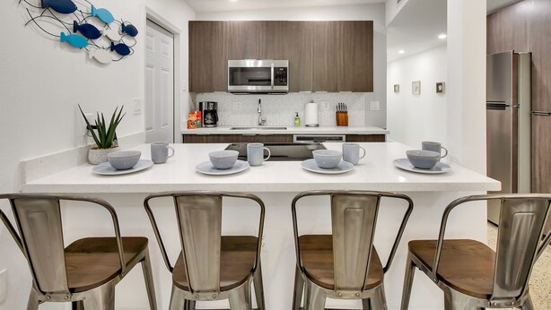 A modern kitchen with white countertops, stainless steel appliances, and wooden cabinets. There are four chairs around a central island with a sink and a coffee maker.