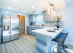 Une cuisine moderne dotée d’appareils électroménagers en acier inoxydable, d’un grand îlot central avec des tabourets de bar et d’une vue sur le jardin à travers une porte ouverte.