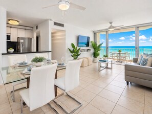 Een moderne woonkamer met een glazen eettafel, witte stoelen, een bank en een balkon met uitzicht op de oceaan.