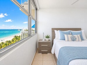 Een slaapkamer met uitzicht op het strand, een bed met blauw beddengoed, een nachtkastje met een lamp en een raam met jaloezieën.