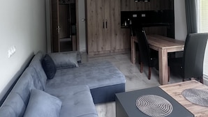 A modern living room with a grey sectional sofa, a wooden dining table, and a kitchen area with wooden cabinets.