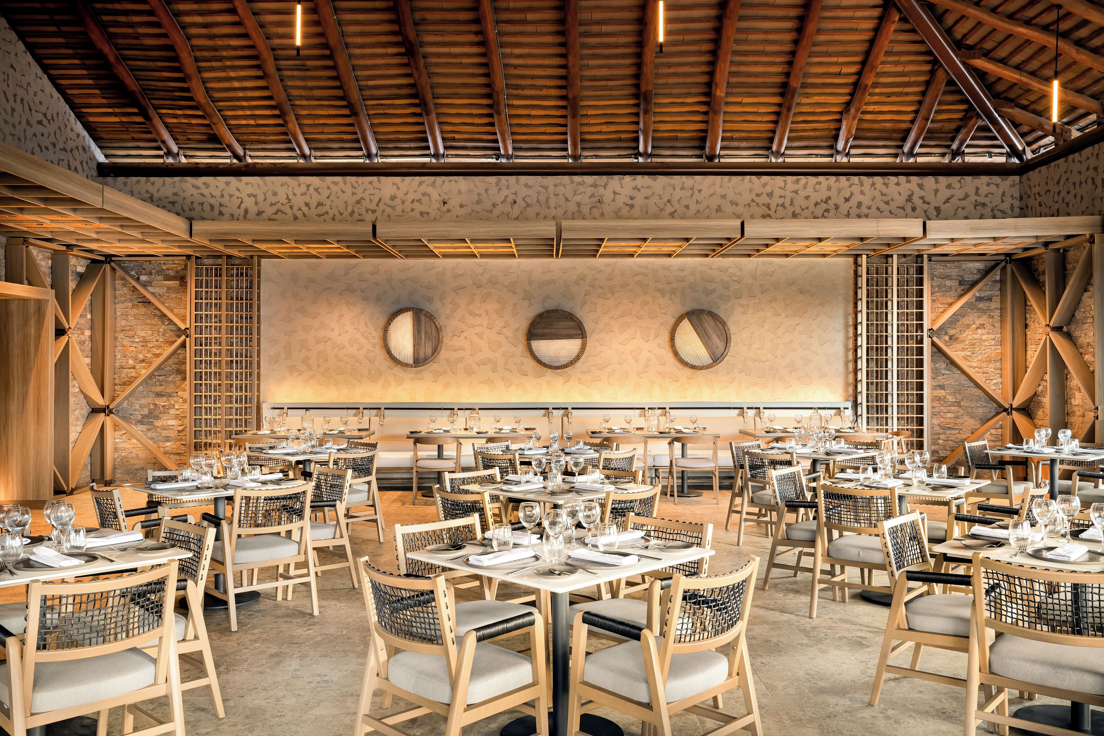 A spacious dining area with multiple tables and chairs, a high ceiling with exposed beams, and a stone wall with circular windows.