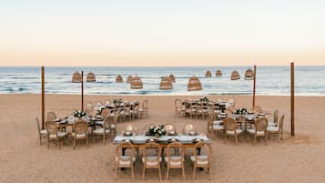 Hochzeitsdekoration am Strand mit runden Tischen und Stühlen, die in einer Halbkreisform zum Meer hin angeordnet sind.
