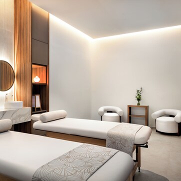 A modern spa room with two massage tables, a wooden panel wall, a round mirror, and a small plant.