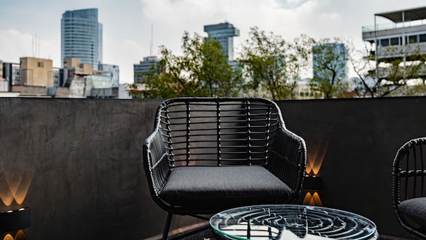 A rooftop terrace with a black wicker chair, a glass table, and a city skyline in the background.