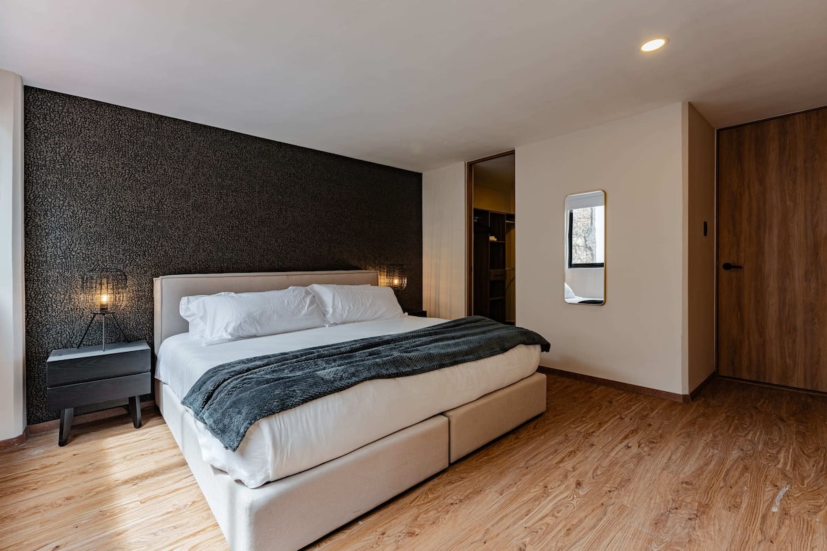 Une chambre moderne avec un grand lit, une table de chevet, une lampe fixée au mur et une armoire en bois.