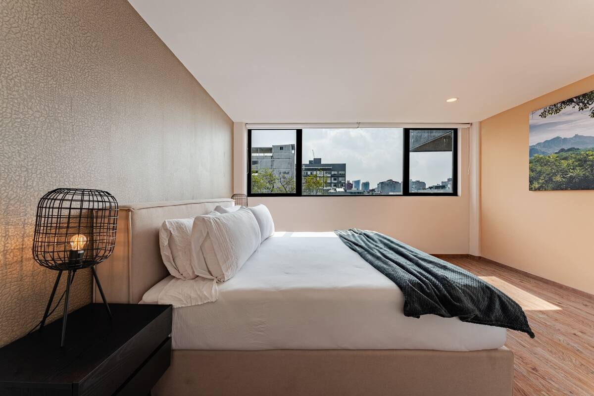 Une chambre moderne dotée d’une grande fenêtre, d’un lit recouvert de linge de maison blanc, d’une table de chevet noire et d’une lampe au design original, faite de fil de métal.