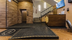 A hotel reception area with a wooden reception desk, a digital screen, a staircase, and a fire extinguisher.