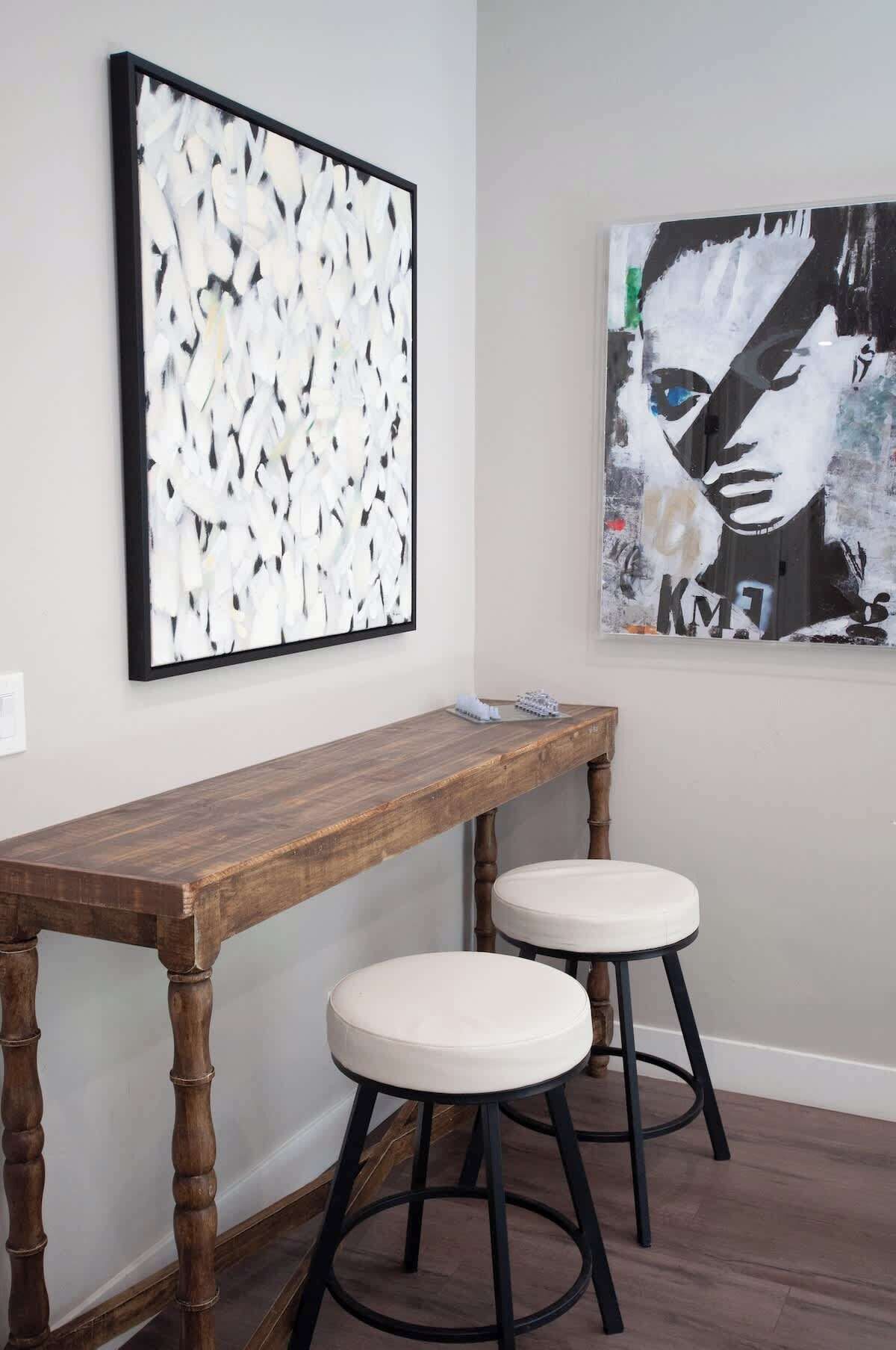 A room with a wooden bench and two stools, a framed abstract painting, and a portrait.