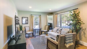 A modern living room with a sofa, coffee table, and TV stand. There are two windows with curtains, a potted plant, and a mirror. - Charming Greensboro Home - 2 Mi to Downtown! (Greensboro)