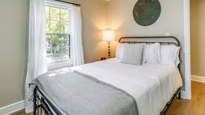A neatly made bed with white linens, a bedside lamp, and a window with white curtains. - Charming Greensboro Home - 2 Mi to Downtown! (Greensboro)