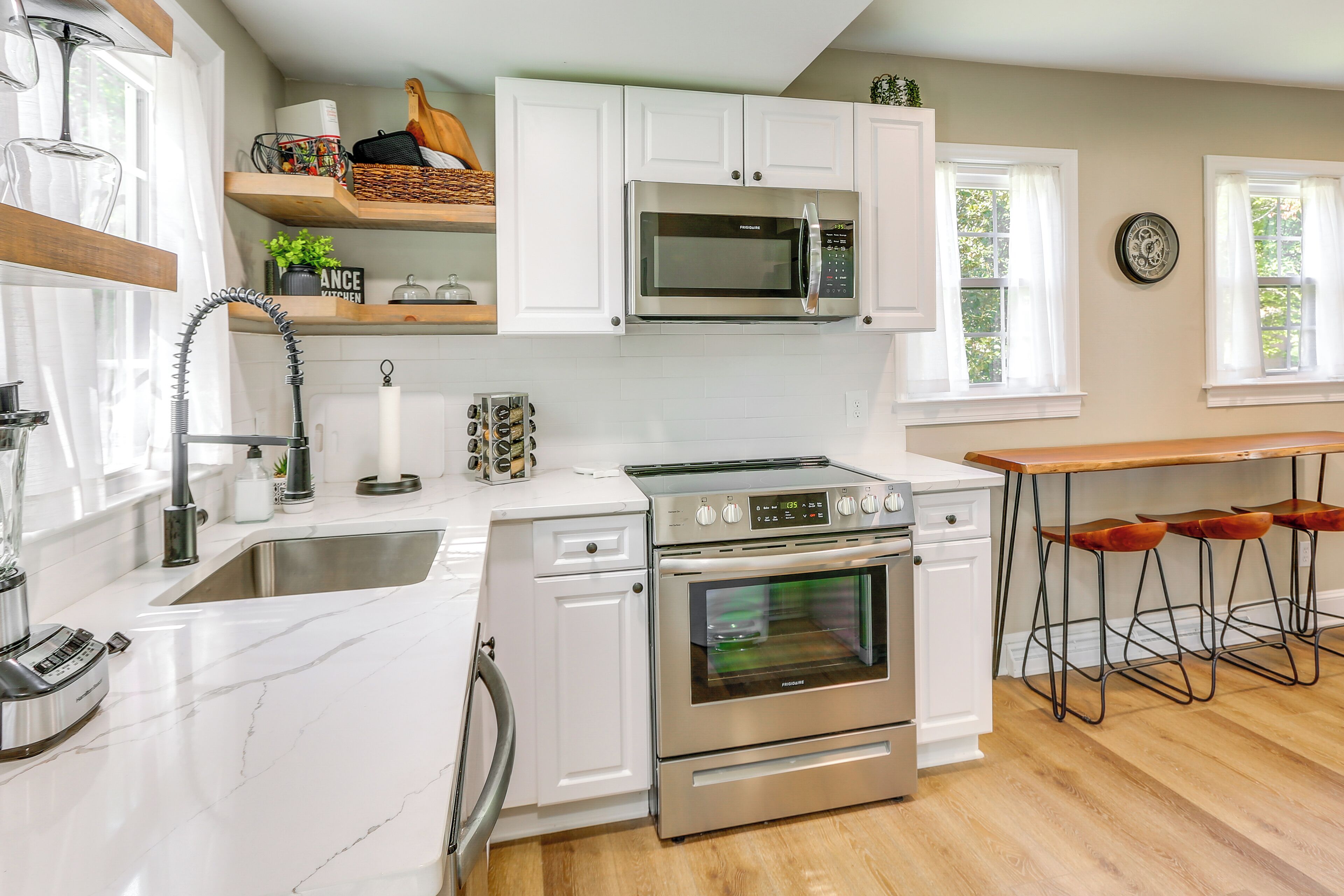 A modern kitchen with white cabinets, a stainless steel microwave, an oven, a sink, and a bar area with stools.