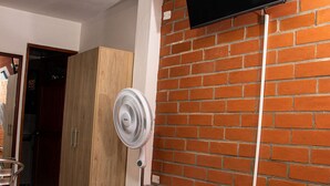 A hotel room with a bed, a TV mounted on a brick wall, a standing fan, and a wooden wardrobe. - Chali (Retiro)