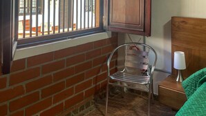 A room with a brick wall, a window with metal bars, a wooden cabinet with a decorative top, a metal chair, a bedside table with a lamp, and a green bedspread. - Chali (Retiro)