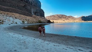A sandy beach with two people walking along the shore, a calm body of water, and rocky hills in the background. - Condo in Crescent Bar Resort—20 minutes from the Gorge amphitheater. (Quincy)
